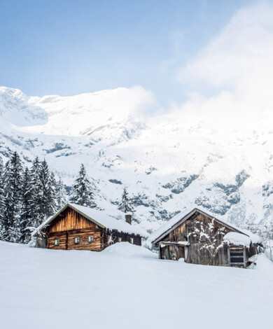 Zwei verschneite Holzhütten vor einer Bergkulisse im Großarltal. 