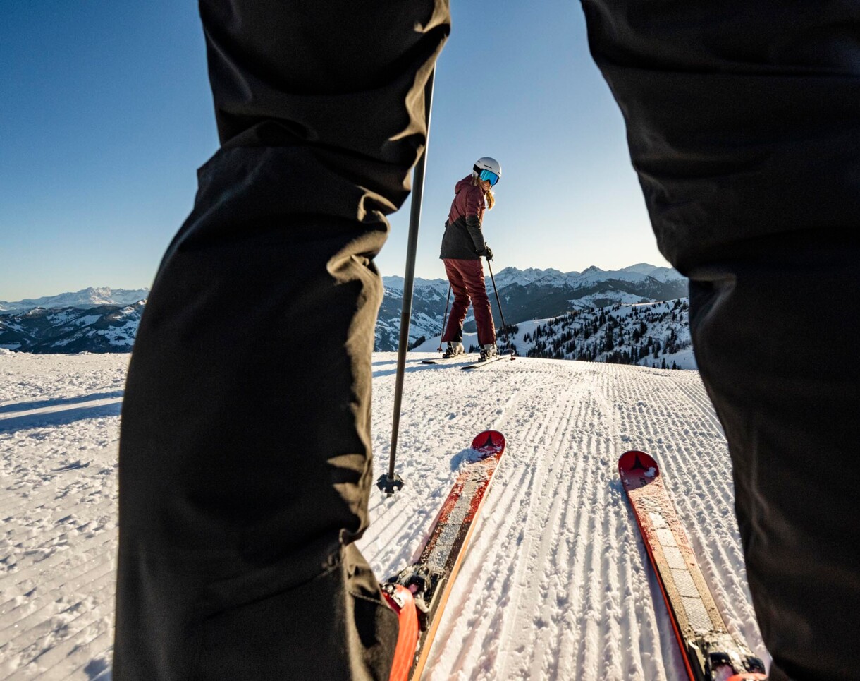 Zwei Skifahrer auf einer verschneiten Skipiste mit Bergblick, aufgenommen aus Perspektive zwischen den Beinen.