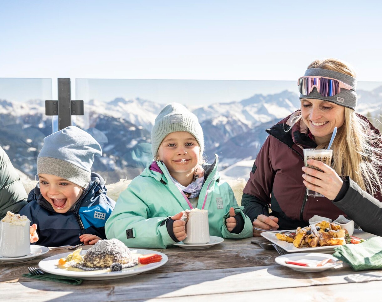 Familie genießt heiße Getränke und Desserts auf einer Terrasse mit Bergblick.
